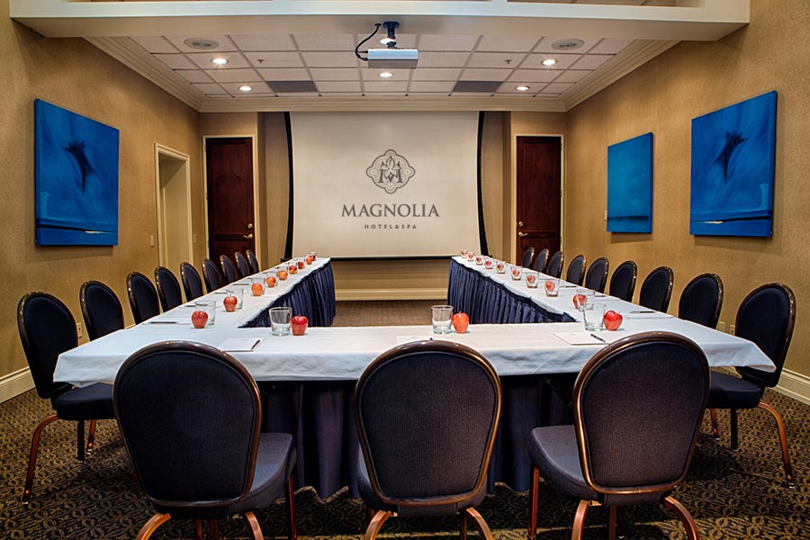 U-shaped meeting room with blue artwork on both walls, projector screen at end of table. Red apples and water settings.