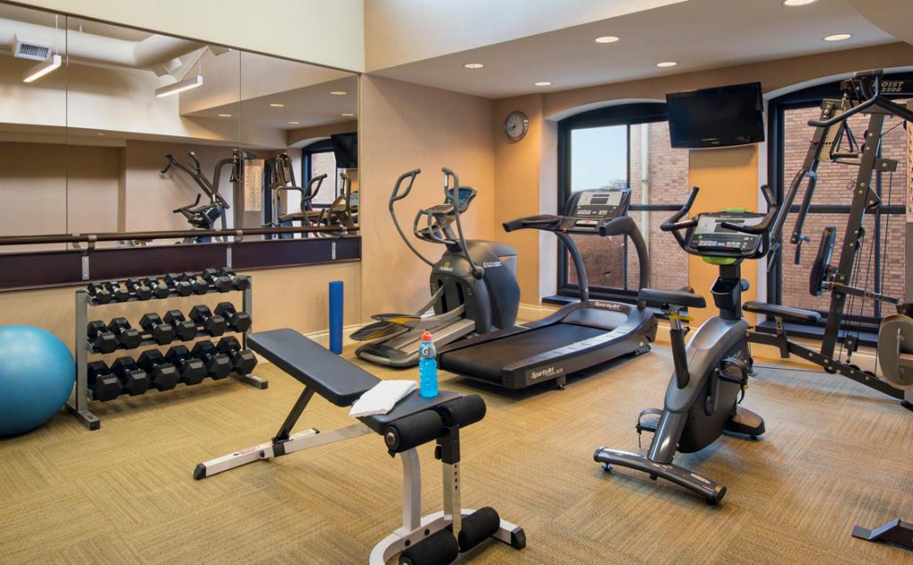 Fitness centre with large windows, weight area, stair master, treadmill, elliptical, and stationary bike. Bottle of blue Gatorade in foreground with towel.