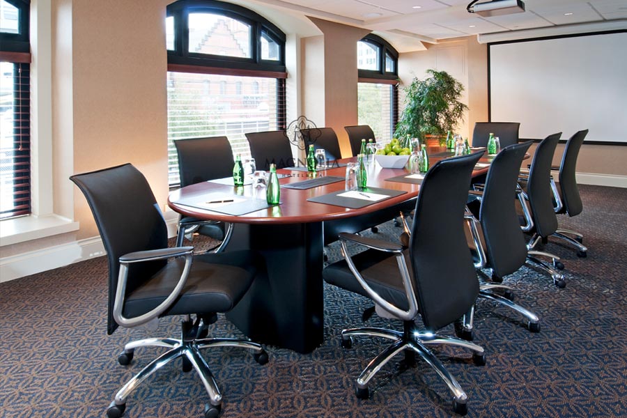 Boardroom table in center of room with natural light from large windows. Projector screen at end of table. Green apples on table with bottles of sparkling water.