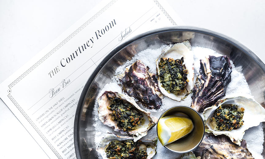 Fresh Oysters in a circle on ice with lemon in centre sitting on top of The Courtney Room Raw Bar and Salads Menu.