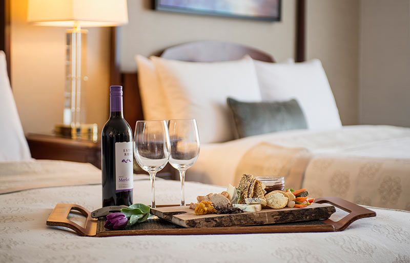 Bottle of red wine with 2 glasses by a selection of fine cheeses, crackers and tasting items on a wood platter on edge of queen sized bed in a room with 2 queen beds.