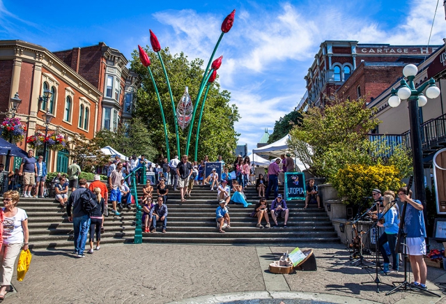 Musicians playing to small crowd of people sitting on steps near open market, blue sky, ornate sculpture, heritage buildings.