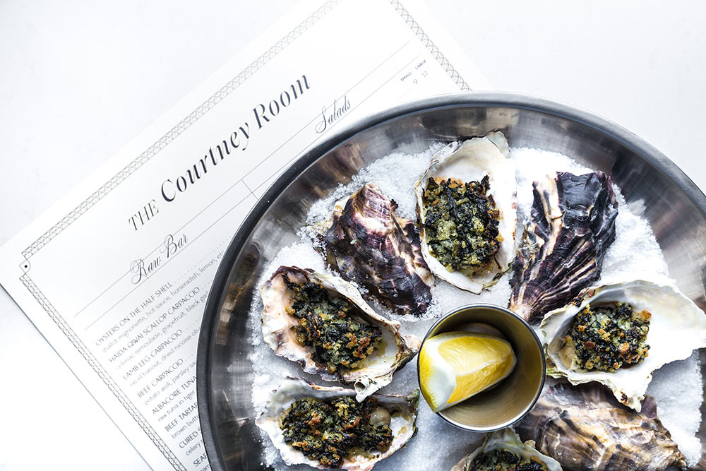 Fresh Oysters in a circle on ice with lemon in centre sitting on top of The Courtney Room Raw Bar and Salads Menu