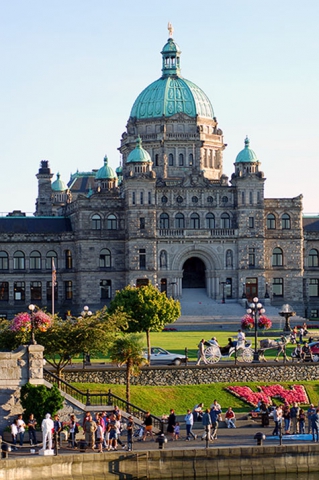 Iconic inner harbour with Parliament building, horse drawn carriage, many people waling along the water.