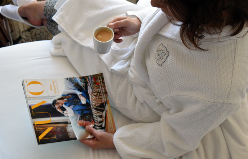 Women drinking Nespresso while reading NUVO magazine in bed, wearing Magnolia embroidered house coat.