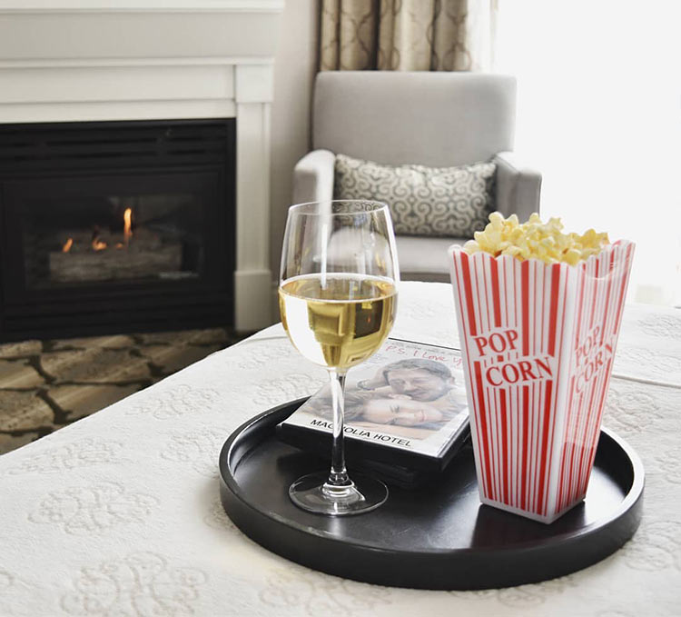 Popcorn, glass of wine and dvd movie on tray on edge of bed with warm fireplace in background