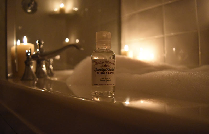 Bottle of hotel size bubble bath with deep soaker tub full of bubbles and candles in behind