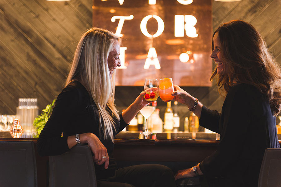 Two women laughing sitting on bar stools cheering with cocktails in hand